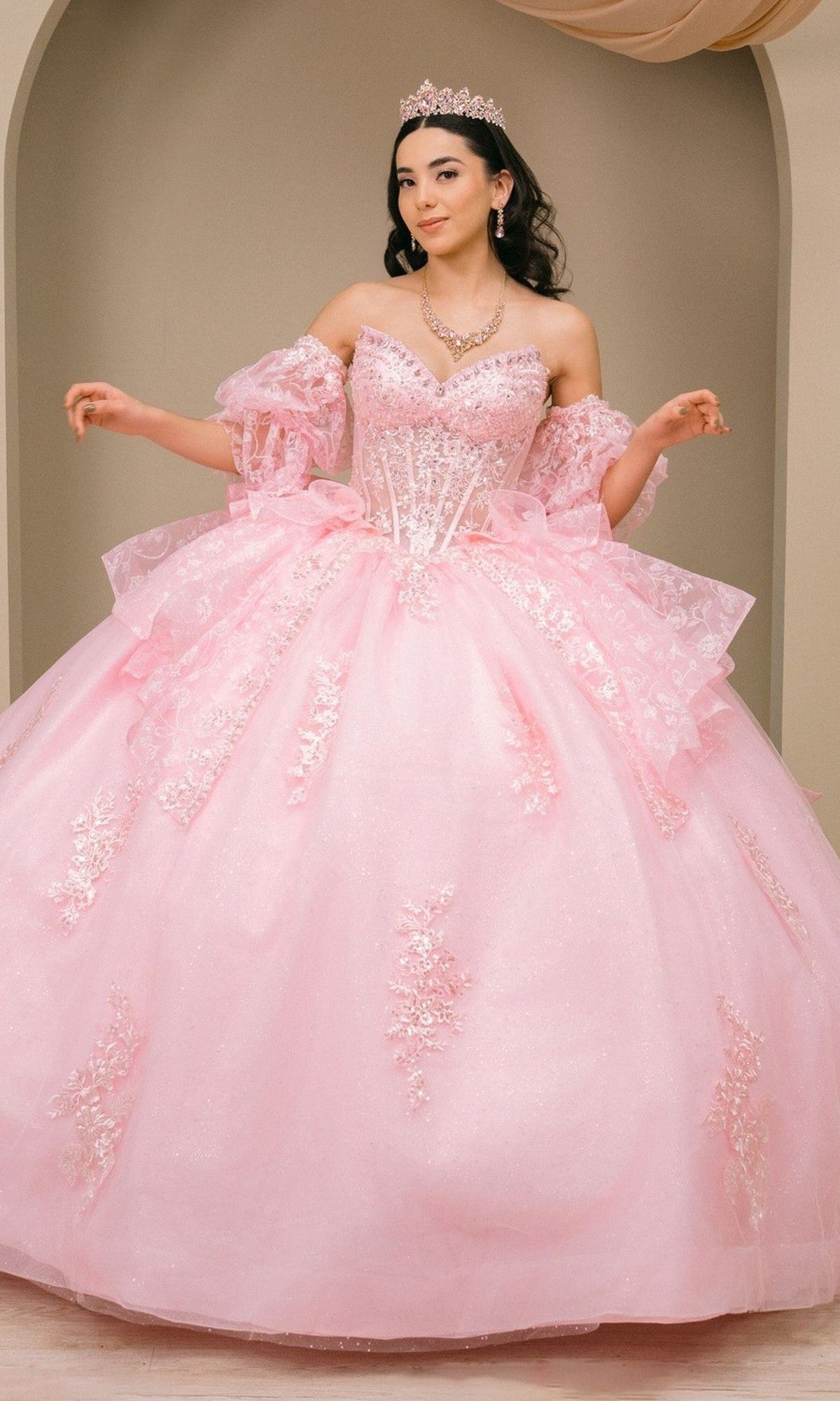 A woman wearing the Quinceanera Dress 8535JQ By Cinderella Couture in pink, featuring floral details and a tiara, stands in front of a beige arched backdrop, posing with her arms slightly raised.